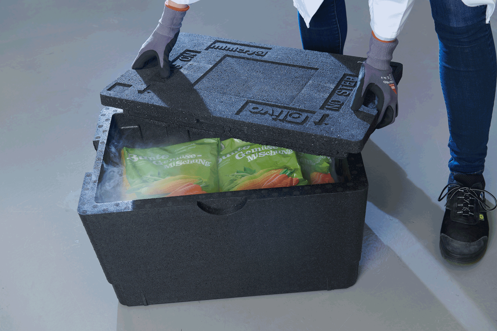 Eine Person mit Handschuhen öffnet einen schwarzen Styroporbehälter mit Beuteln voller gefrorener Karotten. Der Behälter steht auf einem glatten Boden, und um die Öffnung herum ist ein Nebel aus kalter Luft zu sehen.