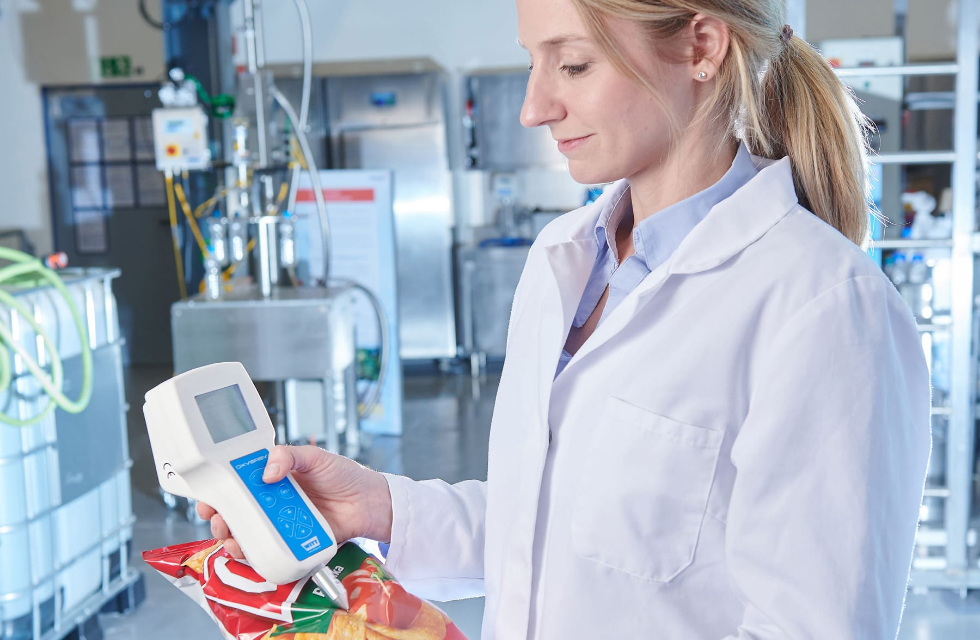 Eine Laborantin in einem weißen Kittel untersucht eine Tüte mit bunten Chips, während sie ein Messgerät in einer Laborumgebung hält.